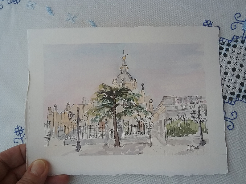 A small watercolor of the Almudena in my hand to see the size. You can see a thumb holding a corner of the watercolor. The watercolor is a line and wash of the Almudena Cathedral with a big evergreen in front of the main gate. You can also see some old time street gas lamps.