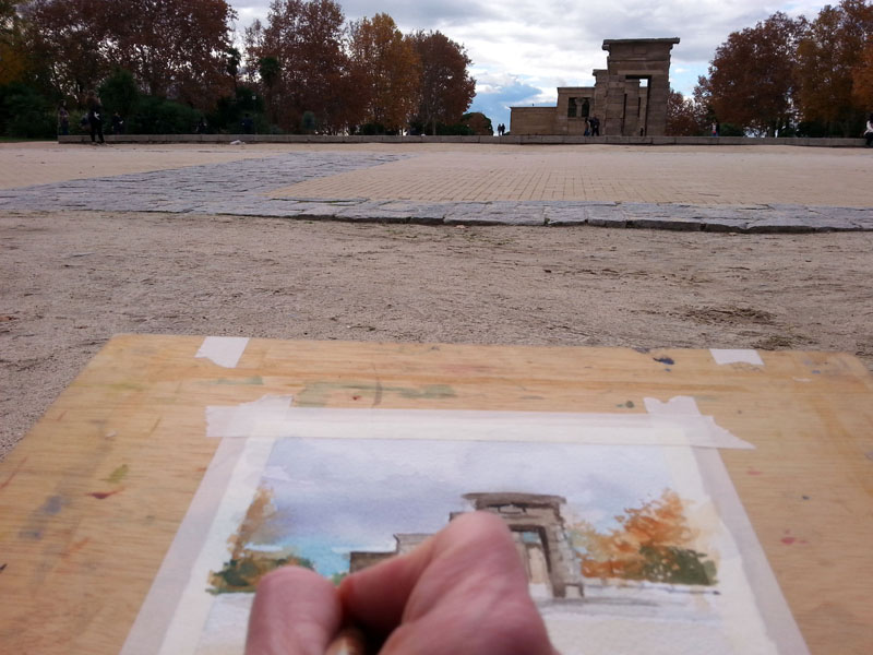This is a picture of a person painting a watercolor of the Temple of Debod. You can see the hand painting and the model which is Debod Temple.