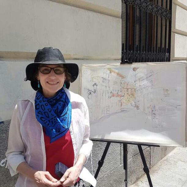Woman standing in front of easel with started watercolor wearing watercolor gear, hat, scarf and long sleeve white shirt and tinted glasses.