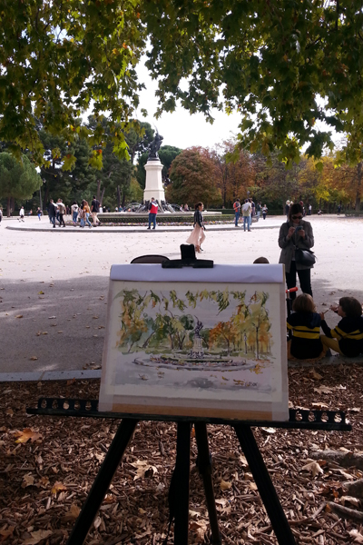 A Watercolor of the Fallen Angel Fountain in the autumn. The watercolor is on an easel and the fountain is in the background